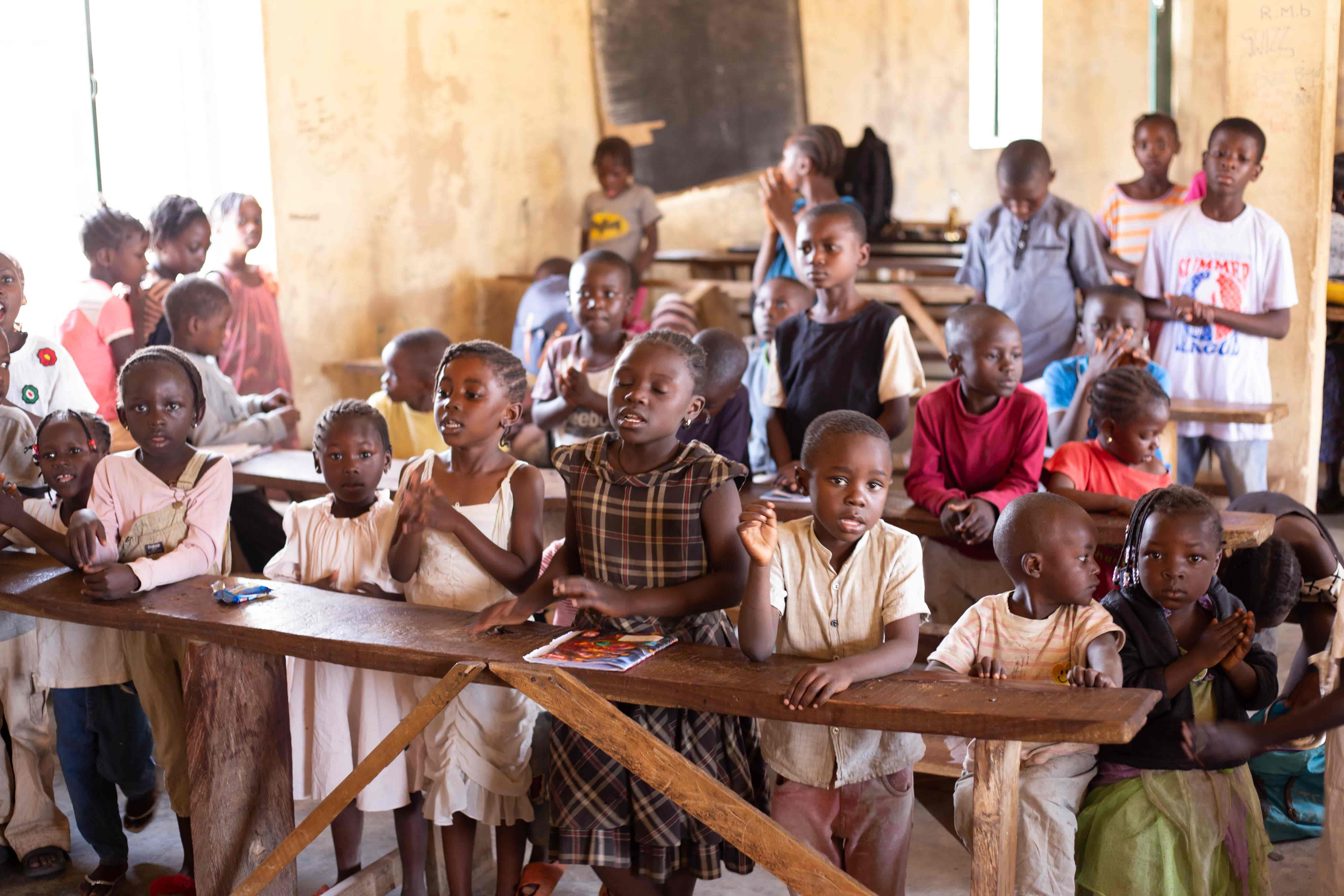 Children standing at wooden desks in a community classroom in Plateau State, singing and clapping together.