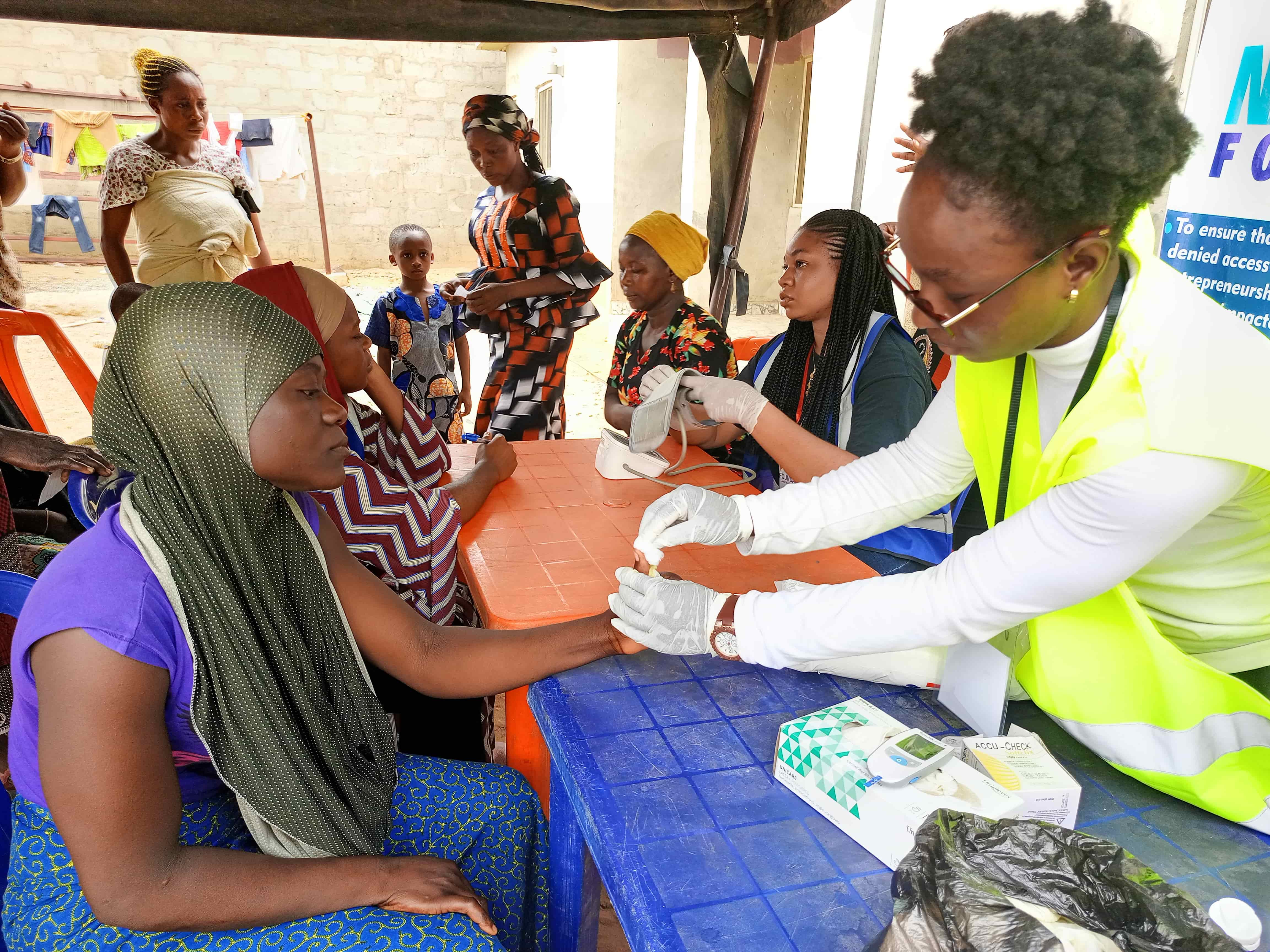 A community health worker in a high-visibility vest performs a finger-prick test for a seated resident during an outdoor outreach session.