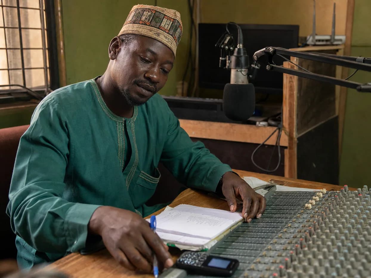 A community radio operator prepares a public health message beside a microphone, mixing desk and basic handset.